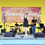 Widok ogólny na scenę amfiteatru na której znajdują wokalistka Kaja Karaplios oraz instrumentaliści. W tle czerwony baner reklamujący Letnie Koncerty. Foto Monika Bilska