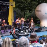 Widok ogólny na scenę i widownię amfiteatru podczas Pikniku Country. Na zdjęciu fragment widowni, scena z muzykami, balon reklamowy oraz motocykl będący dekoracją sceny. Foto Monika Bilska