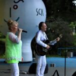 Zbliżenie na sylwetki muzyków zespołu Metrum. Teresa ma na sobie żóltą tunikę i białe spodnie. Ubrany na biało Marek Halup śpiewa i gra na gitarze. Foto Monika Bilska