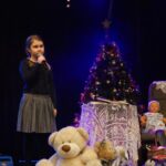 Solistka na scenie z mikrofonem w ręku. Za nią scenografia: choinka, fotel z pledem, stolik z koronkowym obrusem i zabawki dla dzieci. Foto Karolina Gera