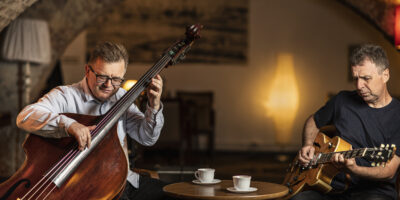 Na zdjęciu muzycy Bohdan Lizoń i Grzegorz Piętak siedzą przy stoliku w kawiarni i grają na instrumentach