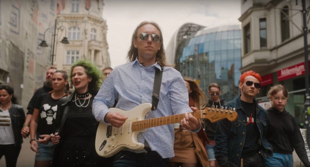 Na zdjęciu widać grupę idących ludzi. Na przodzie ubrany w niebieską koszulę gitarzysta, w okularach przeciwsłonecznych.