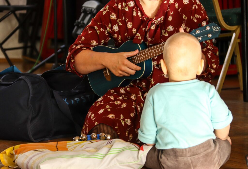 Małe dziecko siedzi tyłęm i słucha jak instruktorka, ubrana w czerwoną sukienkę, gra na małej gitarce